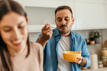 Man eating breakfast in kitchen while woman smiling in foreground