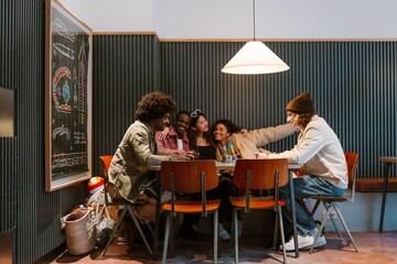 The young multiracial men and women sit around a table in a cozy indoor cafe, smiling, talking, and using a smartphone under warm lighting.