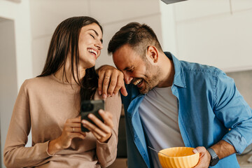 Couple laughing together while reading smartphone in kitchen