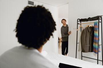 A White adult man smiles while standing by a door, looking at a Black adult man sitting on a bed in...