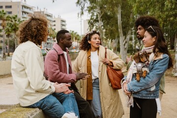 A group of young multiracial men and women stand and sit outdoors in a park near city buildings, talking while one woman holds a small dog.