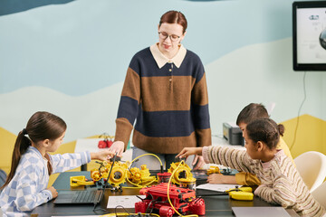 Teacher guiding students in robotic programming class using various educational kits. Children engaged in interactive learning with robotic devices on table