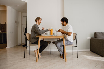 Two adult multiracial men sit at a wooden table and have breakfast at home. One wears a long-sleeve shirt, and the other wears a white t-shirt and pajama pants.