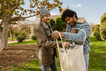 A multiracial gay couple in their 30s stands in a city park on a sunny day, holding a tote bag...