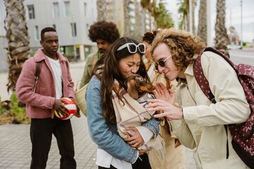 A group of friends in casual outfits walks outdoors, interacting with a small dog. One woman holds the dog, and another man playfully approaches it for a kiss.