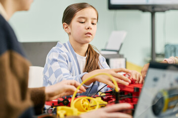 Young girl engaging in STEM activity, assembling components of a robot-building kit on a table alongside classmates within classroom setting