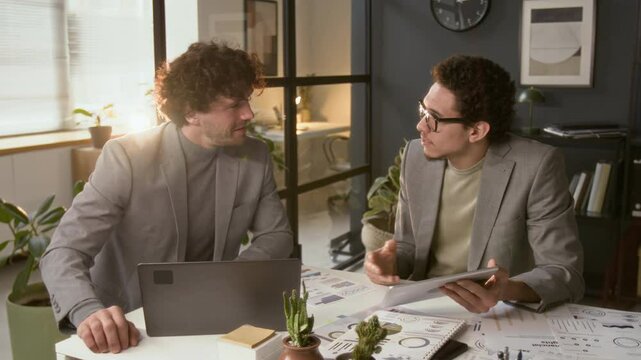 Two young businessmen in formalwear discussing investment opportunities when sitting at office desk with laptop, digital tablet and financial papers