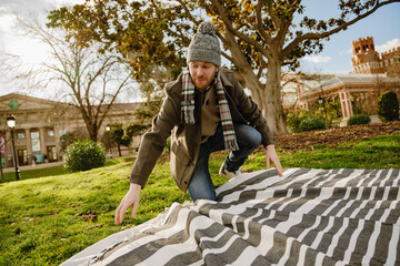 A young White man with a red beard kneeling outdoors, arranging a striped blanket on the grass,...