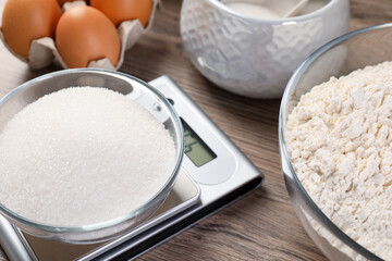 Digital kitchen scale with bowl of sugar and other products on wooden table, closeup