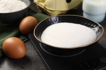 Digital kitchen scale with bowl of sugar and other products on grey table, closeup