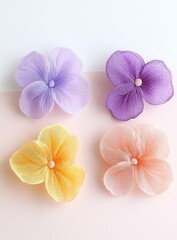 Four delicate, fabric flowers in pastel shades of lavender, purple, yellow, and peach.  Each flower features multiple overlapping petals, a subtle, round center, and soft, flowing textures