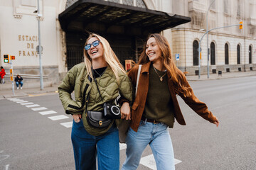 Two women walk arm-in-arm through a city crosswalk, smiling and laughing. They are wearing casual jackets and jeans, one carrying a camera. The background shows Estacio de Franca in Barcelona.