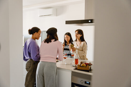 A group of young women stand around a kitchen island at home, eating snacks and drinking wine. They wear sweaters and relaxed clothing in a modern apartment kitchen. - Powered by Adobe