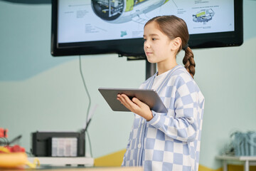 Young girl holding tablet and standing in a classroom with a screen displaying a diagram. She is focused on a tablet, suggesting engagement in a learning activity