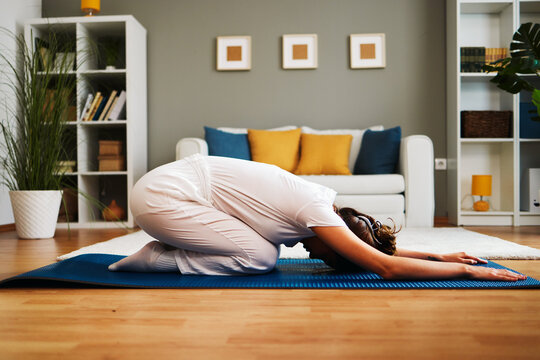 Woman performing balasana yoga pose on mat in living room, promoting relaxation and flexibility