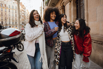 A group of young multiracial women laugh and walk arm in arm on a city street. They wear casual winter clothing, including jackets, scarves, and a leopard coat.
