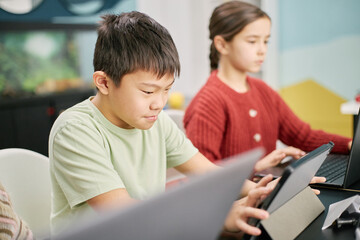 Diverse students engaged in classroom activities using tablets for learning, one student focuses intently on screen while classmate in background follows along
