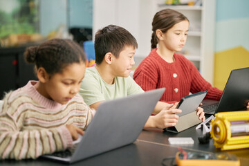 Group of children using laptops and tablets in classroom setting, focusing intently on screens, demonstrating learning through technology