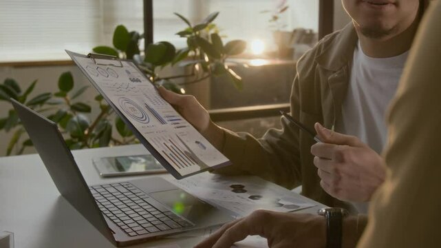 Midsection view of young male employee discussing statistical data with unknown coworker while holding clipboard with document and pointing at graphics with pen