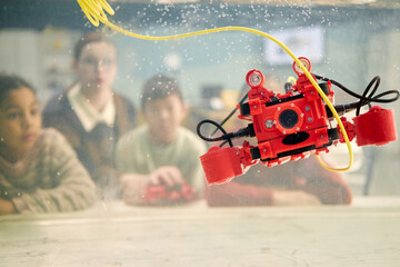 Students in classroom eagerly observing red underwater drone submerged in aquarium. Background showing blurry students engaged in learning about underwater technology