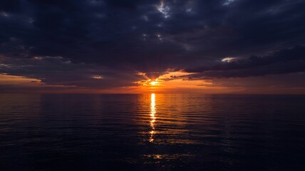 Obraz premium Dramatic sunset over calm ocean waters with sun rays piercing th