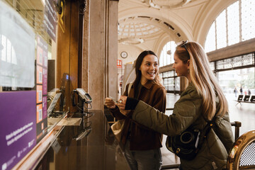 A woman stands and smiles while holding tickets and looking at her female friend standing next to her
