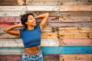 Portrait cheerful woman poses playfully with her hands behind her head, smiling against wooden wall
