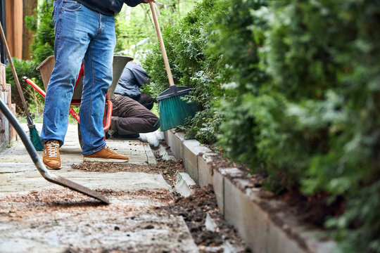 A gardener working in a summer garden, sweeping a path with a broom and cleaning up soil from plants