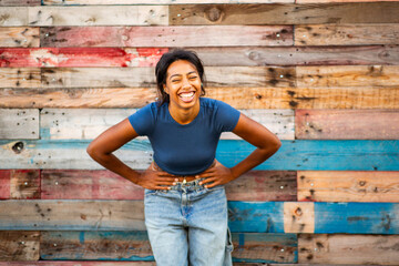 cheerful young woman with hands on her hips, laughing in front of colorful wooden wall.