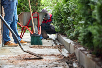 A gardener working in a summer garden, sweeping a path with a broom and cleaning up soil from plants