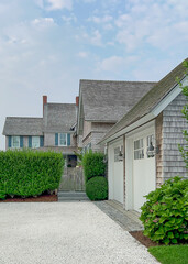 A beautifully maintained coastal home with cedar shingles, a gravel driveway, white double garage doors, lush greenery, and a bright blue sky dotted with soft clouds.