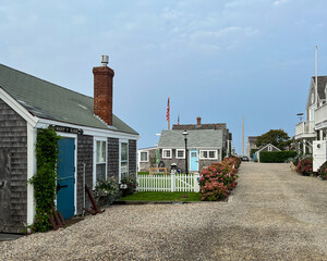 A picturesque coastal street featuring cedar-shingled cottages, a bright blue door, a white picket fence, blooming hydrangeas, and an American flag under a serene, clouded sky.