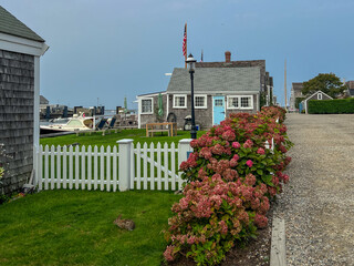 A charming coastal scene with a white picket fence, vibrant hydrangeas, a cedar-shingled cottage with a blue door, a boat dock, and a small rabbit resting on lush green grass under a calm sky.