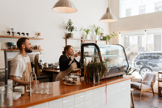 A male employee stands behind the bar and looks at the female owner who puts a plate on the display, they smile, in a cafe