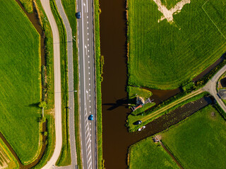 Aerial view of a Dutch countryside with a traditional windmill by a canal, green fields, and a nearby road with moving cars under a clear blue sky.