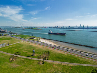 Aerial view of cargo ships navigating a wide river or channel near an industrial port with cranes and smokestacks in the background under a clear blue sky.