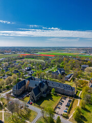 Aerial view of a peaceful suburban neighborhood with tree-lined streets, modern and traditional buildings, and surrounding farmland stretching into the horizon under a clear sky.