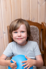 Blue-eyed boy blowing up a blue balloon while sitting on a wooden chair on a wooden background