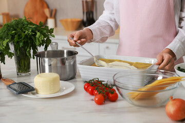 Woman spreading bechamel sauce onto spinach lasagna at marble table indoors, closeup