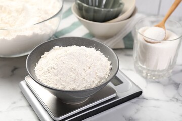 Digital kitchen scale with bowl of flour and other products on white marble table, closeup