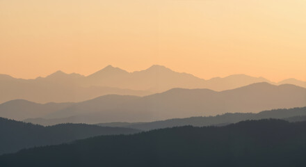 Obraz premium Mountain ranges in layers at sunset with golden orange sky and silhouetted pine forest in foreground. Tranquil natural landscape for travel destinations