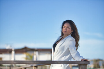 Young, beautiful, brown-haired woman in a white jacket, looking at the camera, relaxed and calm, leaning on a wooden railing.