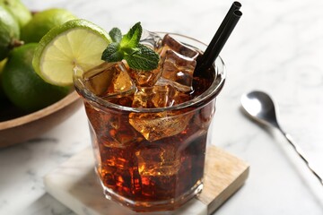 Tasty Cuba Libre cocktail in glass, limes and spoon on white marble table, closeup