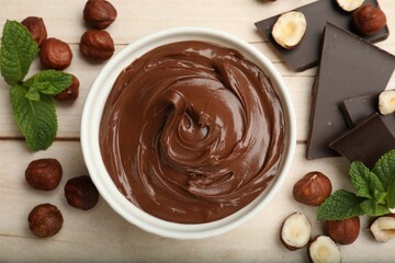 Chocolate hazelnut spread in bowl, nuts, pieces of chocolate and mint on wooden table, flat lay