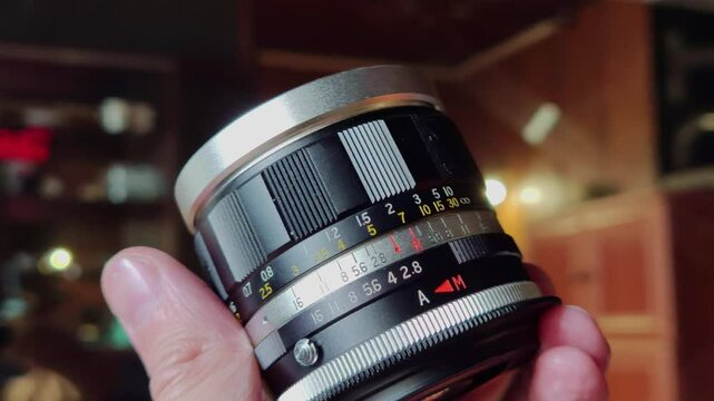 A close-up shot of a hand carefully holding a vintage camera lens. The lens features intricate markings for focus distance and aperture settings, with a silver top ring and a black and white body
