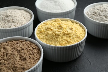 Different types of flour in bowls on black textured table, closeup