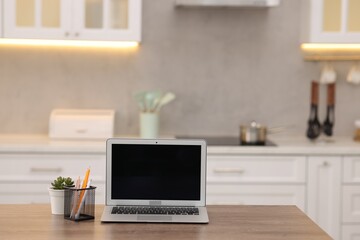 Laptop with blank screen, houseplant and stationery on wooden table in kitchen. Mockup for design