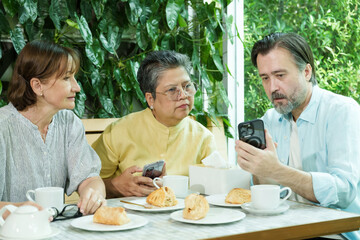 Asian senior woman and two Caucasian friends focus on a smartphone screen while sitting at a cafe table, enjoying pastries and tea in a joyful and relaxed retirement atmosphere.