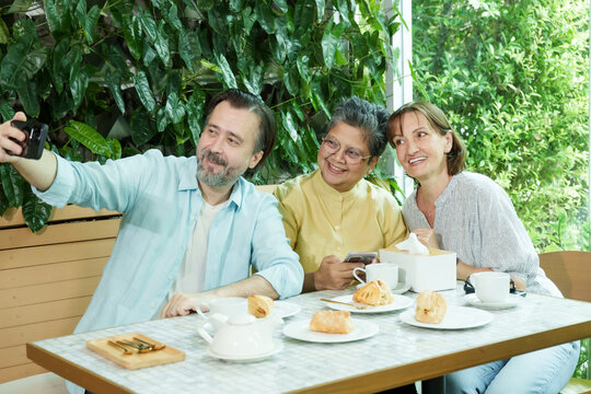 Asian senior woman and two Caucasian friends laugh and pose for a selfie at a cafe table, surrounded by pastries and tea, enjoying a joyful and relaxed retirement afternoon together. - Powered by Adobe
