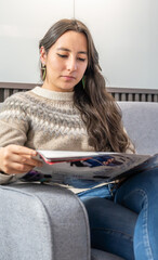 A young woman is sitting comfortably on a modern grey sofa in what appears to be a clinic or office waiting room with a magazine
